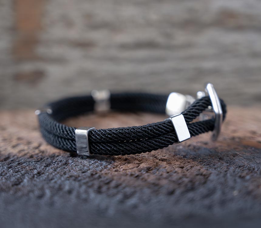 Right side view of the Maris Sal Nautical Anchored New Haven bracelet in black resting on top of a piece of rustic wood
