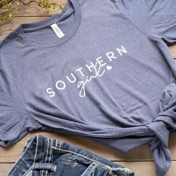 Flat lay of the Humm & Willow Southern Girl women's graphic tee on a wood top with jean shorts and sandals