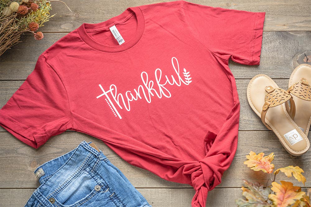 Flat lay of the Humm & Willow Thankful women's graphic tee on a wood top with jean shorts and sandals