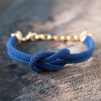 Front view of the Maris Sale Nautical nautical knot bracelet with navy rope and gold chain and charm on a rustic wooden top