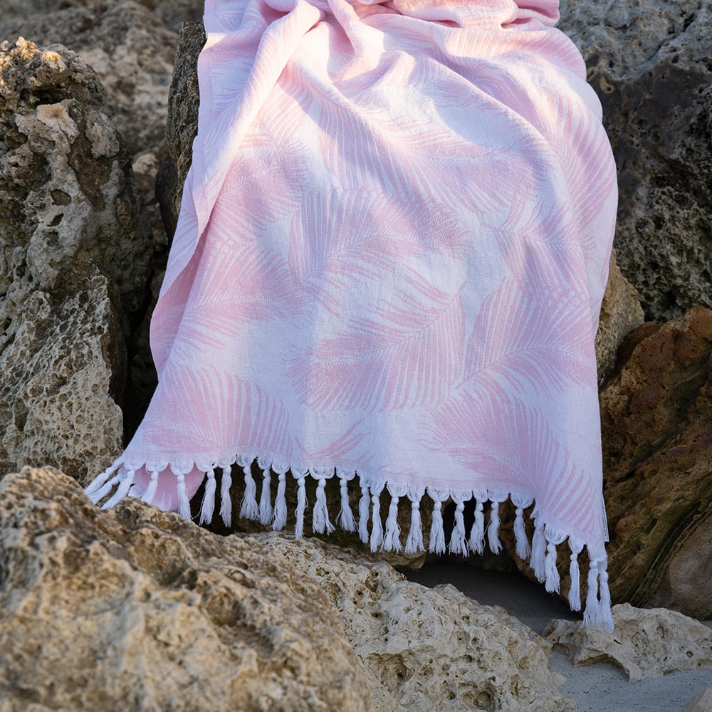 Pink Palms Turkish Towel by Case+Drift draped over rocks along a shoreline