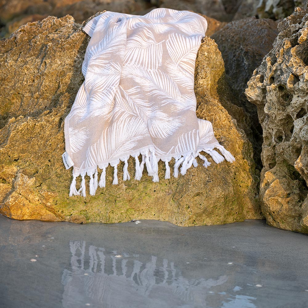 Tan Palms Turkish Towel by Case+Drift draped over rocks along a shoreline