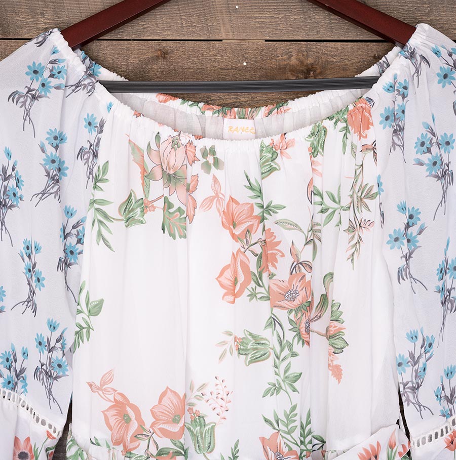 Closeup View of the Top of the Ranee’s Floral Off-Shoulder Dress Hanging Against a Wood Backdrop