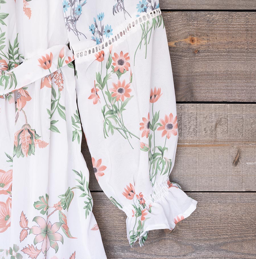 Closeup view of the sleeve of Ranee's floral off-shoulder dress hanging on a hanger against a a wood background