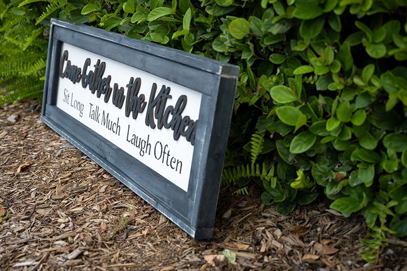 Angled view of the Come Gather in the Kitchen wood sign resting along shrubs