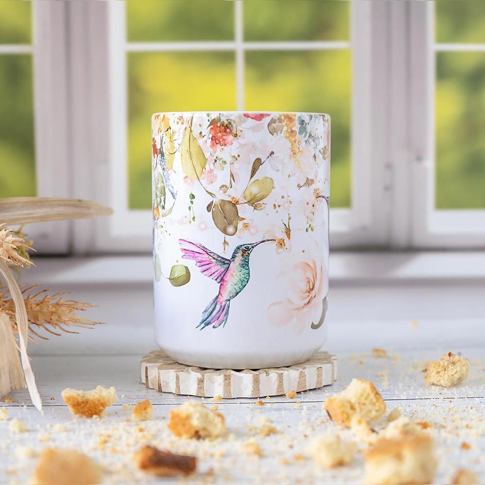 Front view of the Florae & Snow Hummingbird coffee mug against a window and on a coaster and wood top