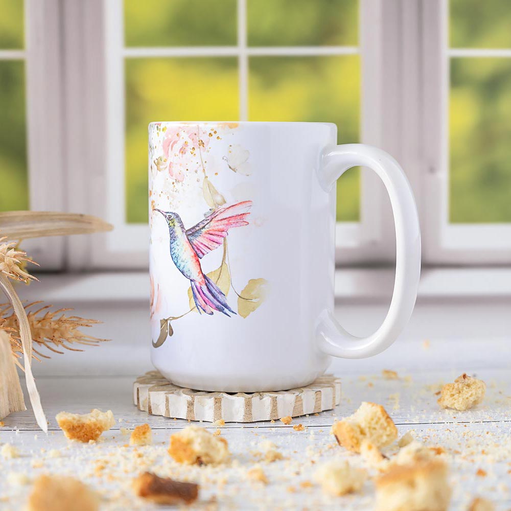 Left angle view of the Florae & Snow Hummingbird coffee mug against a window and on a coaster and wood top