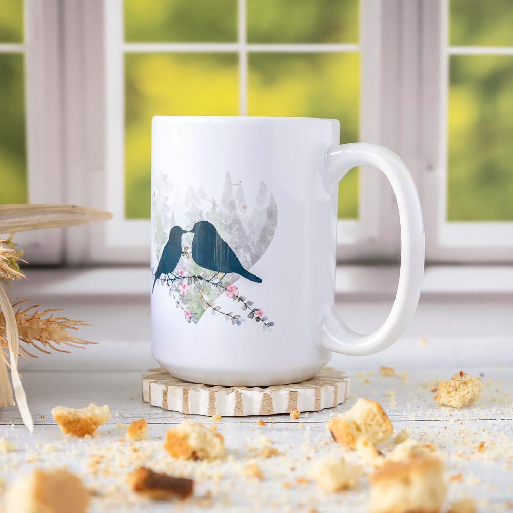 Left angle of the Florae & Snow Lovebirds Day Mug series of two lovebirds in front of a heart on th emug on wooden top layered with biscotti and in front of a window