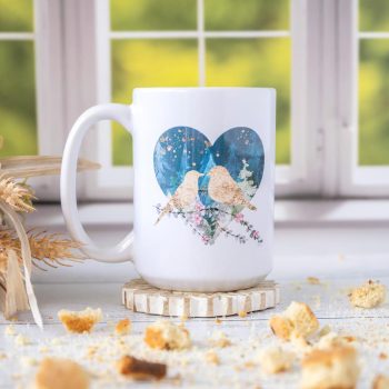 Right view of the Florae & Snow Lovebirds Night Mug series of two lovebirds in front of a heart on th emug on wooden top layered with biscotti and in front of a window