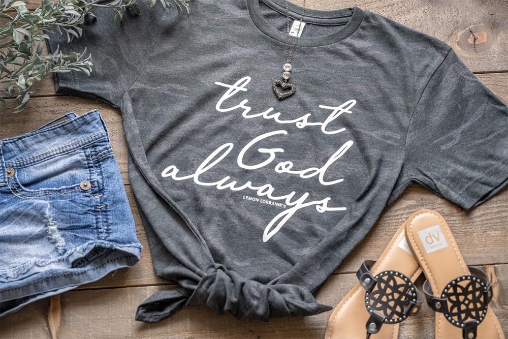 Flat Lay of the women's trust god always graphic tee on a wooden top with jean shorts and sandals