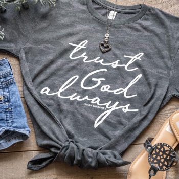 Flat Lay of the women's trust god always graphic tee on a wooden top with jean shorts and sandals