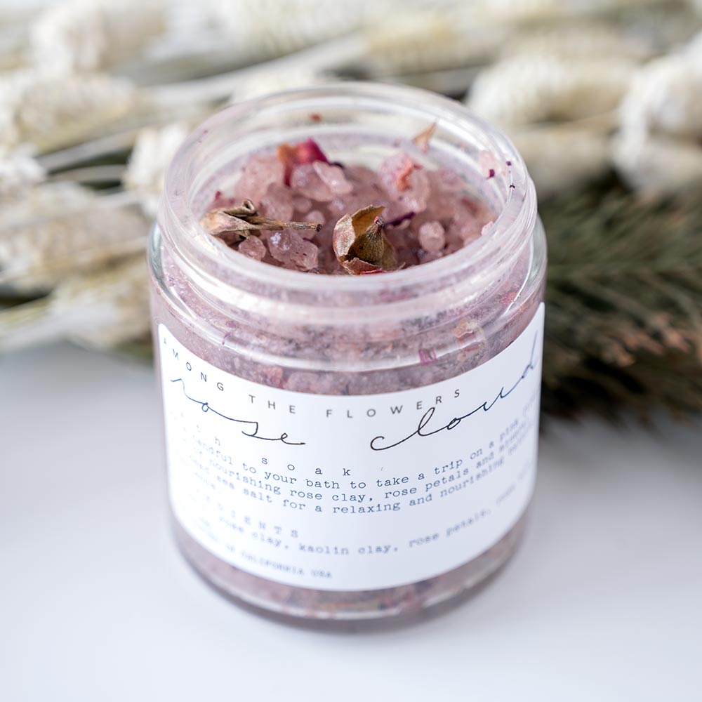 Among the Flowers Rose Cloud Bath Soak with Open Jar Looking at the Salts with a Rustic Background with Flowers and Greenery