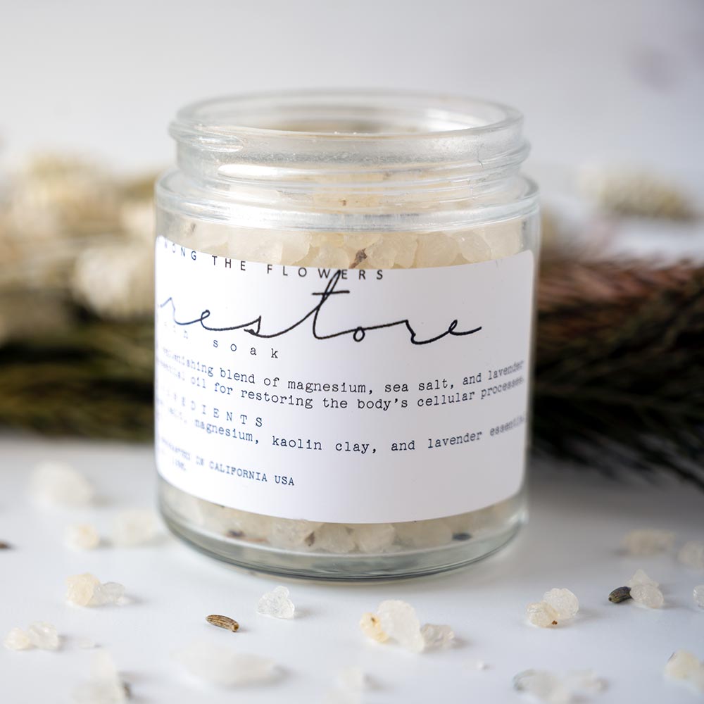 Among the Flowers Restore Bath Soak with Open Jar and Salts Poured out with a Rustic Background with Flowers and Greenery
