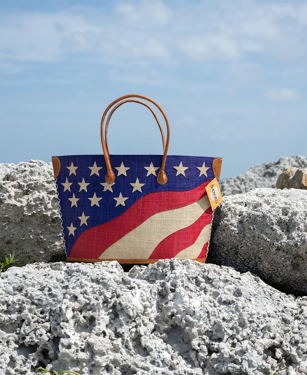Front view of the Shebobo American Flag Straw Tote Bag at the beach on rocks