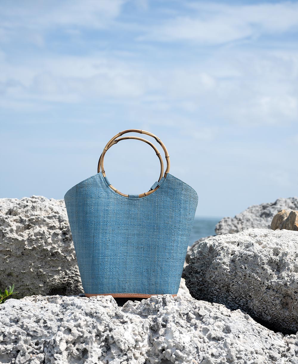 Back view of the Shebobo Carmen Straw Bucket Basket bag in turquoise on the rocks at the beach