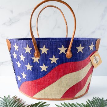 Front view of the Shebobo American Flag Straw Tote Bag against a clean white and marble background with palm leaves in front
