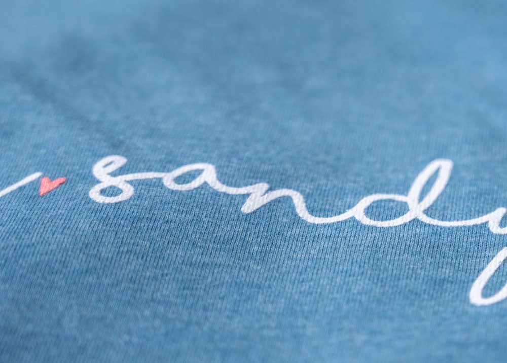 Closeup of the typography, printing and fabric of the Salty. Sandy. Happy women's graphic tee on a wooden boardwalk background with nautical and beach accents around it
