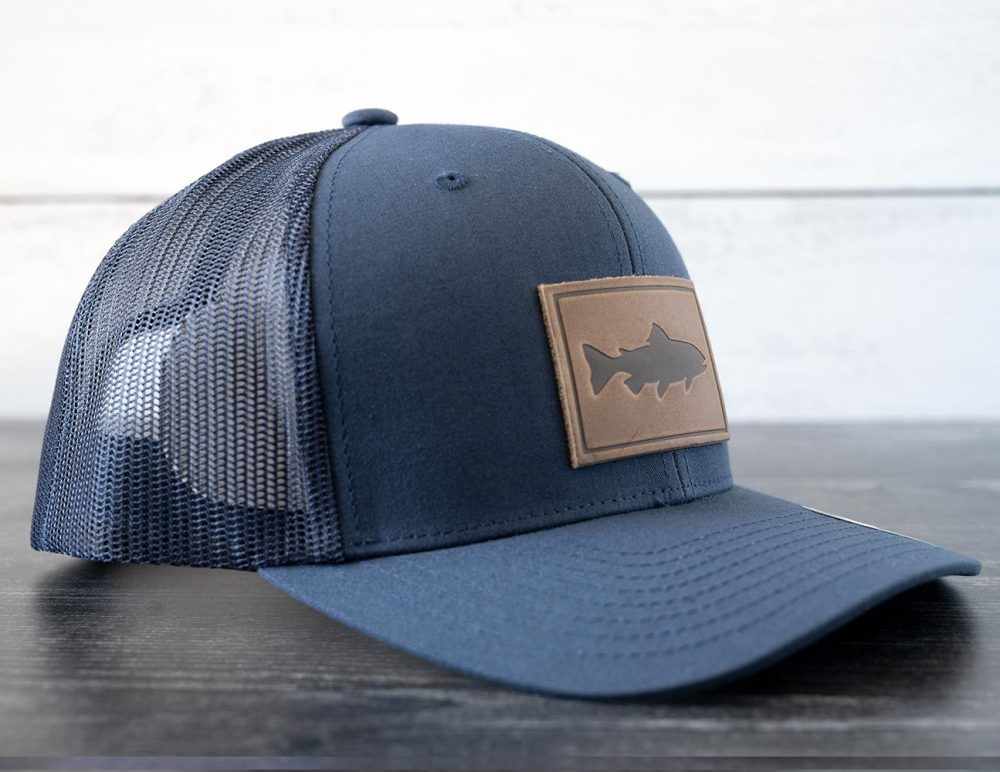 Left angled view of the RANGE Leather Trout hat in the color navy against a white and black rustic wood background