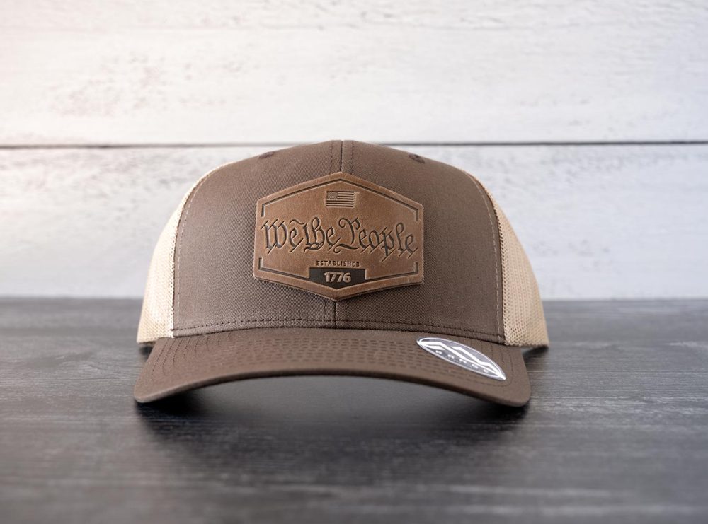 Front view of the RANGE Leather We the People hat in the color brown & khaki against a white and black rustic wood background