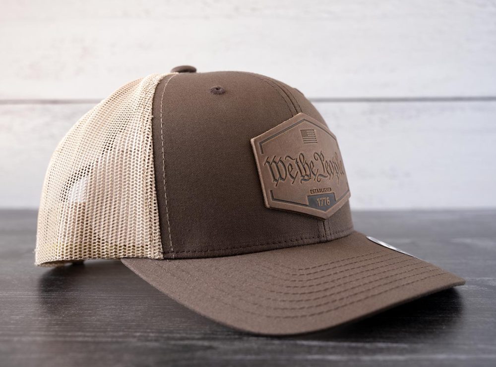 Left angled view of the RANGE Leather We the People hat in the color brown & khaki against a white and black rustic wood background