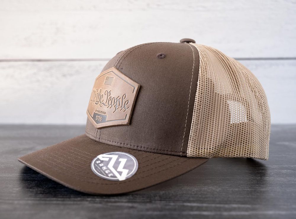 Right angled view of the RANGE Leather We the People hat in the color brown & khaki against a white and black rustic wood background