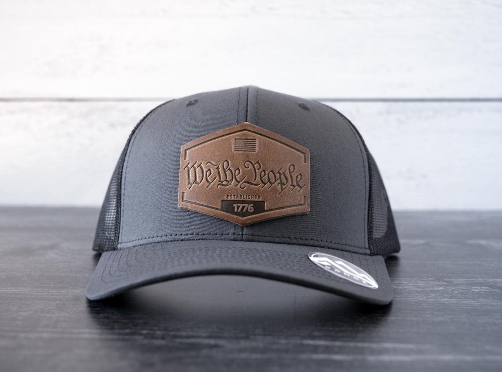 Front view of the RANGE Leather We the People hat in the color charcoal against a white and black rustic wood background