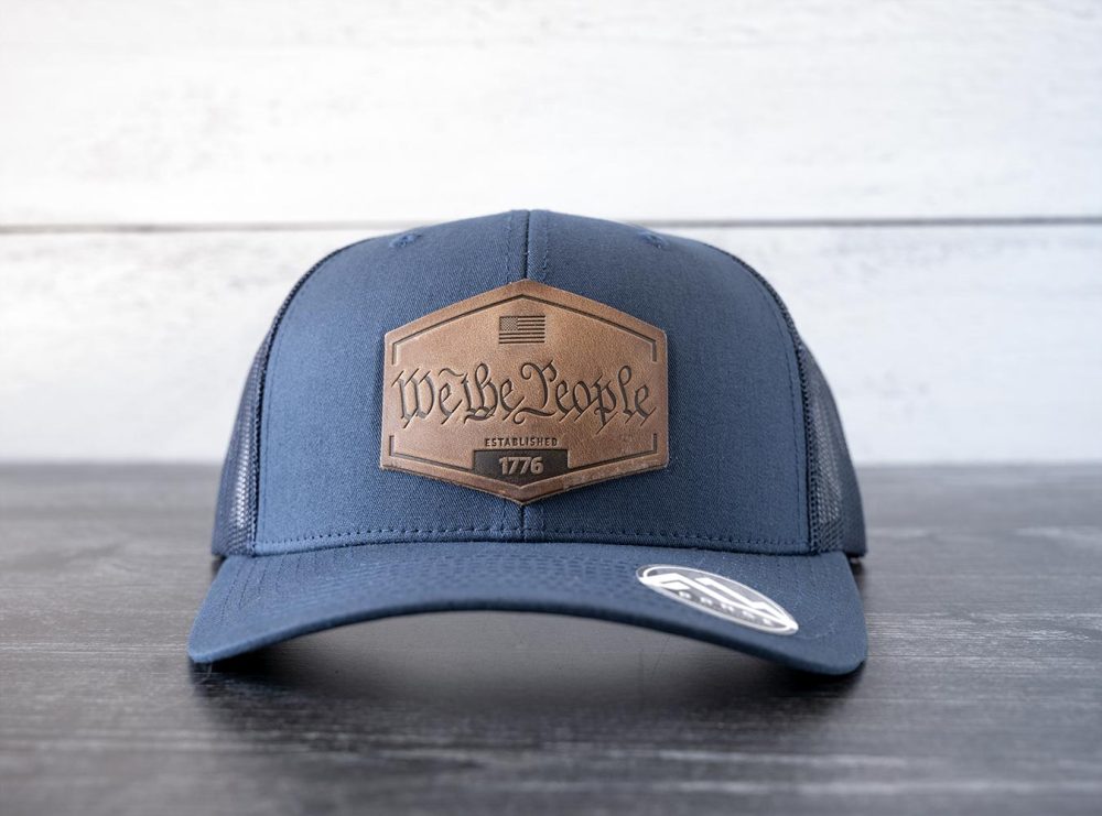 Front view of the RANGE Leather We the People hat in the color navy against a white and black rustic wood background