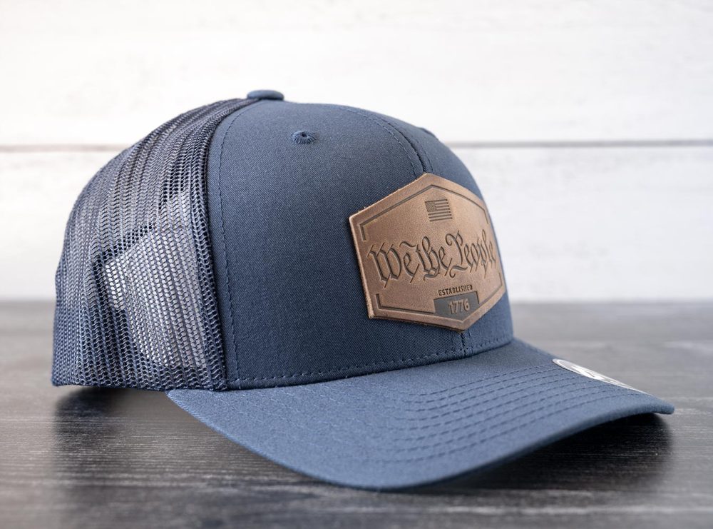 Left angled view of the RANGE Leather We the People hat in the color navy against a white and black rustic wood background