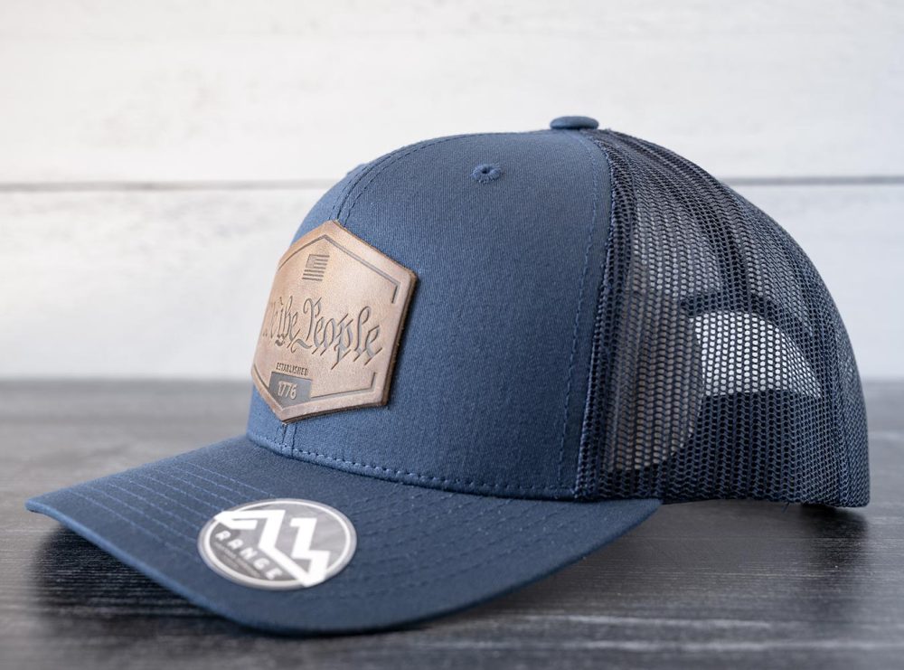 Right angled view of the RANGE Leather We the People hat in the color navy against a white and black rustic wood background