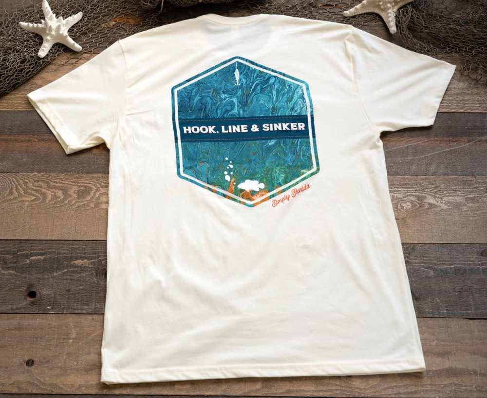 Back view of the SImply Florida Hook, Line & Sinker graphic tee resting against a rustic wood background and nautical background accents of fishing net